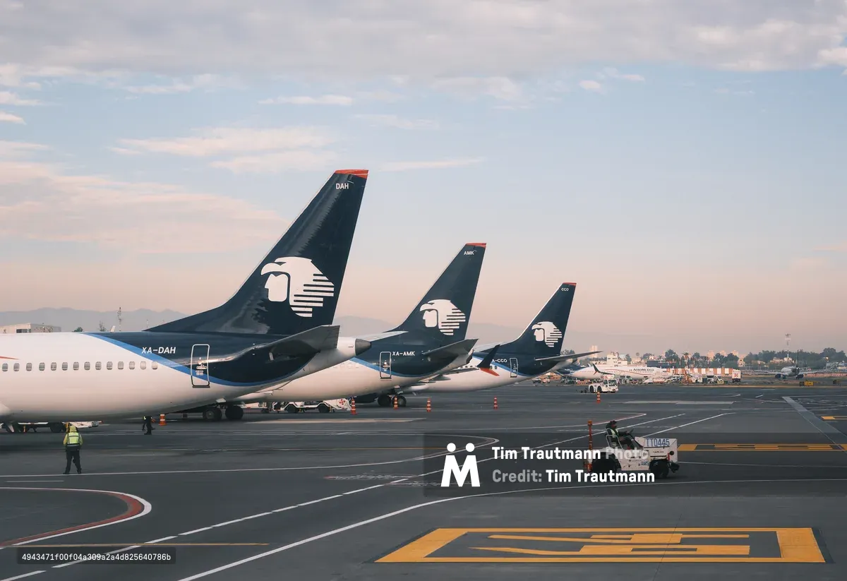 Three Aeroméxico passenger jets are parked in a row on the tarmac at Mexico City International Airport (Aeropuerto Internacional Benito Juárez), their dark navy tail fins bearing the airline's distinctive Aztec eagle logo receding in perspective from left to right. The aircraft registration numbers XA-DAH, XA-AMK, and XA-CCO are visible on the fuselages. A ground crew worker in a high-visibility vest walks near the closest aircraft, and a small white tug vehicle operates in the foreground. Orange traffic cones and yellow tarmac markings are visible on the dark pavement. Additional aircraft from other airlines are parked in the distance. The sky is pale blue with soft pink-tinted clouds, suggesting early morning or late afternoon light.