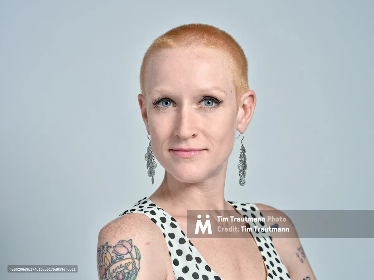 Cayce Ryan presents a confident, contemporary portrait against a clean studio backdrop in Portland's industrial Southeast district. Her platinum buzz cut frames piercing blue eyes enhanced with subtle makeup, while vintage-inspired drop earrings complement her polka-dotted halter top. Colorful tattoo artwork adorns her shoulders, creating visual interest against the minimalist composition and soft, even lighting that emphasizes her professional yet artistic persona.