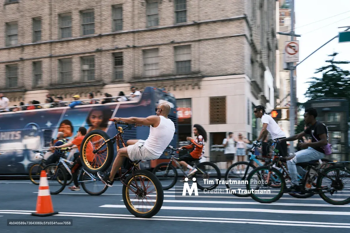 A dynamic street scene captures the kinetic energy of Manhattan's cycling culture as riders navigate the crosswalk at 7th Avenue. The central figure, a bald man in a white tank top, executes a dramatic wheelie on his bicycle with orange-rimmed wheels, his body language expressing pure urban swagger. Behind him, a blue city bus creates a striking backdrop against the weathered brick facades of Midtown buildings, while other cyclists and pedestrians blur through the frame, suggesting the constant motion of New York street life. The golden hour light bathes the scene in warm tones, transforming an ordinary intersection into a stage for spontaneous urban theater.