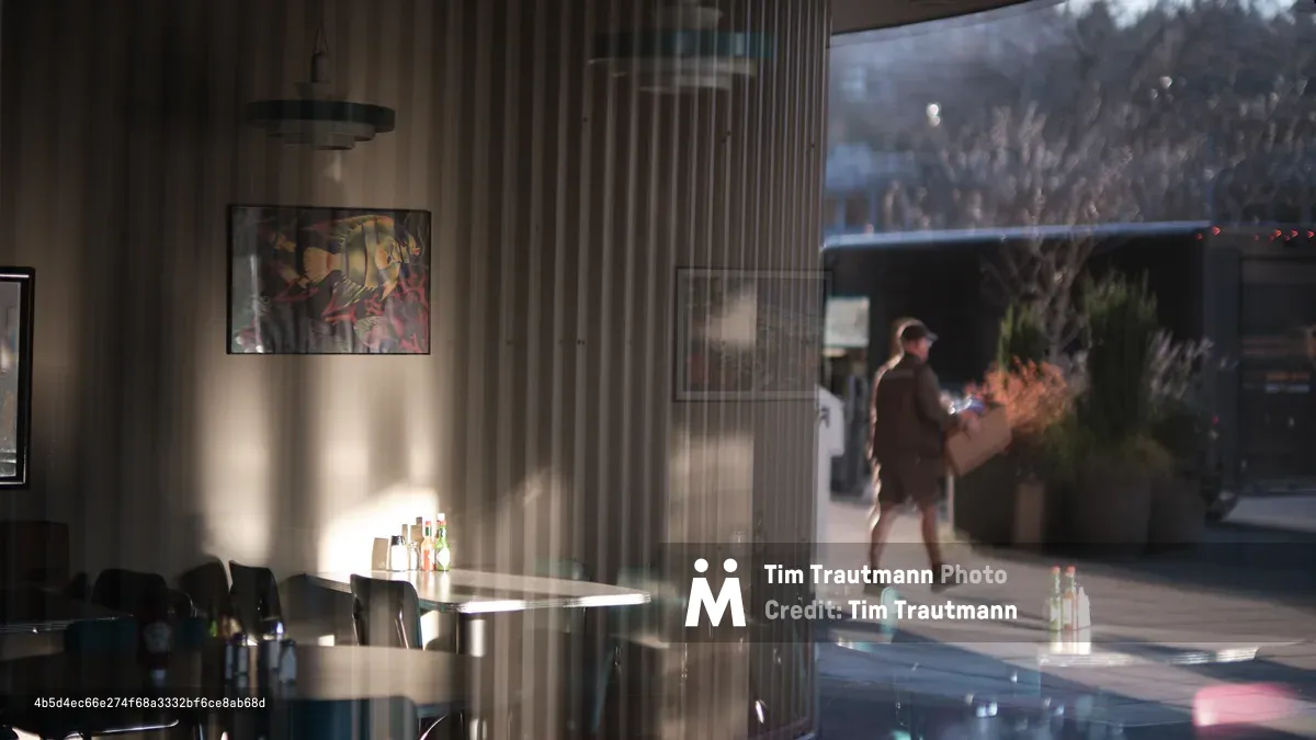 Warm afternoon light streams through floor-to-ceiling windows of a modern Seattle café, creating dramatic shadows and reflections across polished surfaces. A solitary figure moves through the sun-dappled space while geometric patterns from window frames dance across cream-colored walls adorned with contemporary artwork. The interplay of natural light and architectural elements transforms the intimate dining space into a study of light, shadow, and urban tranquility.