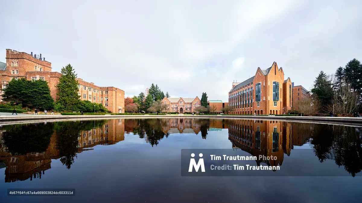 A perfectly still reflecting pool mirrors the iconic red brick architecture of the University of Washington campus under an overcast Pacific Northwest sky. The scene captures both historic collegiate Gothic buildings on the left and the modern angular forms of newer academic structures on the right, their warm terracotta facades creating a harmonious architectural dialogue. Towering evergreens frame the composition while scattered students traverse the distant pathways, adding human scale to this serene academic sanctuary. The glassy water surface doubles the visual impact, creating a symmetrical dreamscape where sky and earth converge.