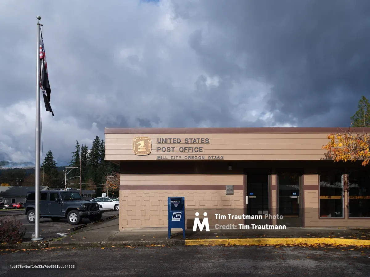 The United States Post Office in Mill City, Oregon stands resolute beneath a dramatic canopy of steel-gray clouds that threaten rain. The modest brick and siding building, marked by its official postal service signage and the zip code 97360, anchors this small Pacific Northwest community against a backdrop of towering evergreens. An American flag hangs limp in the still air while a blue USPS collection box stands sentinel beside the entrance, and autumn leaves scatter across the wet pavement, suggesting the changing season in this Cascade foothills town.