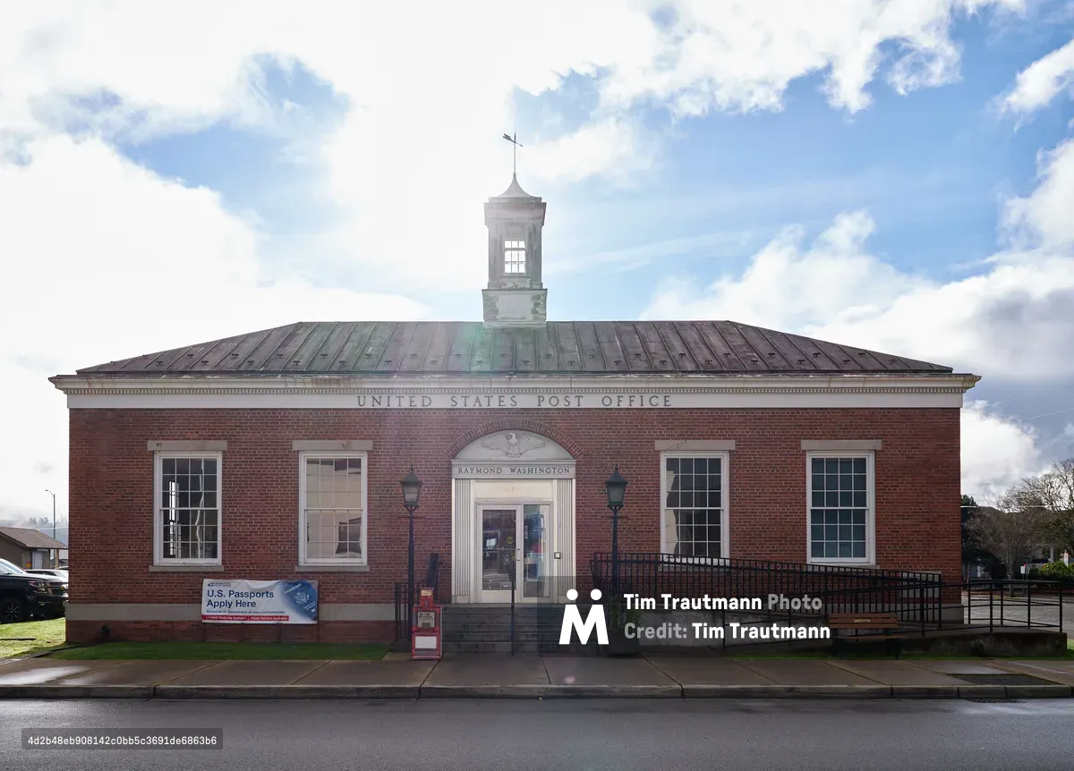 A stately red brick United States Post Office stands proudly under a dramatic sky of billowing white and gray clouds in Raymond, Washington. The symmetrical Georgian Revival architecture features white-trimmed windows flanking a central arched entrance, topped by a distinctive octagonal cupola with weathervane. The weathered copper roof catches subtle green patina against the moody Pacific Northwest sky, while wet pavement reflects the building's imposing presence on Duryea Street.