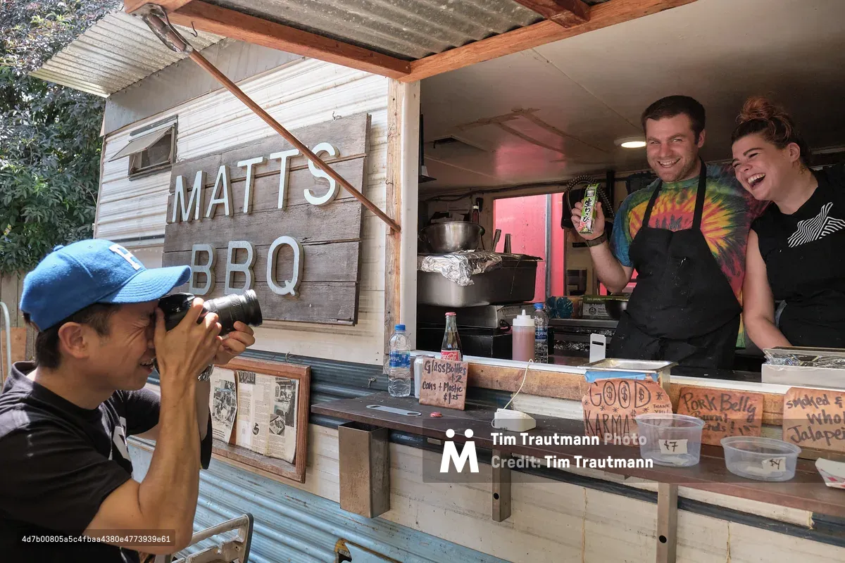A Japanese tourist wearing a blue baseball cap photographs two laughing staff members at Matt's BBQ food cart on Mississippi Avenue in Portland, Oregon. The male staff member in a tie-dye shirt and black apron holds up a small Japanese snack item for the camera, while his female colleague beside him laughs. The rustic wood-sided cart displays a hand-lettered sign reading "Matt's BBQ," along with menu signs for pork belly and smoked jalapeño. Handwritten signs on the counter read "Good Karma" and "Glass Bottles $2."