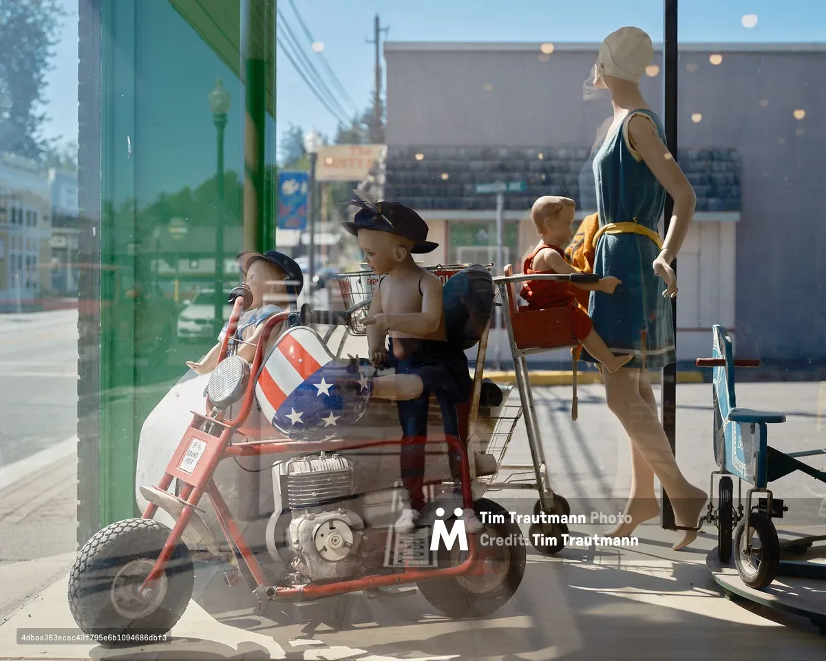 A quirky antique or thrift store window display in Vernonia, Oregon, photographed through the glass with the sunny small-town street reflected in it. The display features several vintage child mannequins posed on and around a red mini motorcycle with an American flag-patterned fairing. A female adult mannequin in a teal 1920s-style dress and white cap pushes a vintage metal shopping cart with a child mannequin seated in it. A small vintage tricycle sits to the right. The main street of Vernonia, including a green lamppost, utility poles, and storefronts, is visible in the reflection.