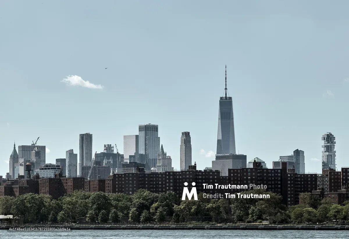 The gleaming spire of One World Trade Center pierces a hazy afternoon sky, dominating Manhattan's Financial District skyline as viewed across the Hudson River. Red-brick housing projects stretch along the waterfront in the foreground, their uniform facades creating a textural contrast against the soaring glass and steel towers beyond. Lush summer foliage softens the urban landscape while a solitary bird glides through the pale blue expanse above the city's architectural symphony.