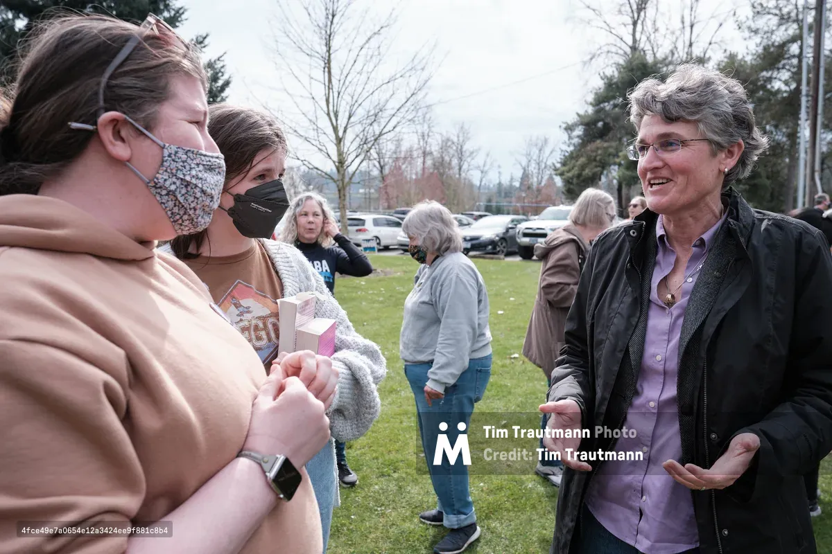 An outdoor community event in Oregon City, Oregon showing people engaged in conversation on a grassy area with cars parked in the background. Two women on the left wear face masks while speaking with an older woman in glasses and a dark jacket on the right, with other attendees visible throughout the scene.