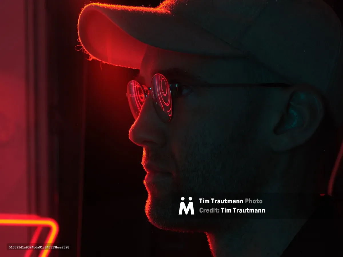 A dramatic side profile portrait of a person wearing reflective sunglasses and a hat, illuminated by vibrant red and blue neon lighting in a dark urban setting. The neon light creates striking rim lighting effects and colorful reflections in the eyewear.