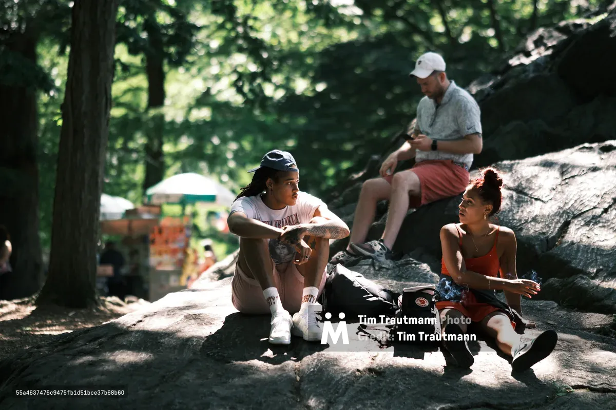 Three friends find solace on the weathered schist outcroppings of Central Park, their casual conversation punctuated by dappled sunlight filtering through the canopy above. A young woman in a black cap and white tee sits cross-legged in contemplative repose, while her companion in an orange tank top leans back in easy conversation. Behind them, a man in a baseball cap checks his phone, perched on the ancient Manhattan bedrock that forms the park's natural amphitheater.