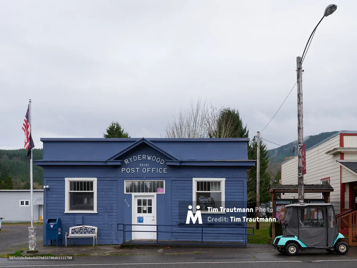 A weathered blue clapboard post office stands as the community anchor in rural Ryderwood, Washington, its white-trimmed windows and peaked roofline bearing the dignified simplicity of small-town America. The overcast Pacific Northwest sky casts a muted light across the scene, while an American flag flutters beside the modest building and a turquoise utility vehicle waits nearby. Forested hills rise in the background, framing this intimate portrait of postal service in logging country. The composition captures both the isolation and enduring civic pride of this unincorporated Cowlitz County community.