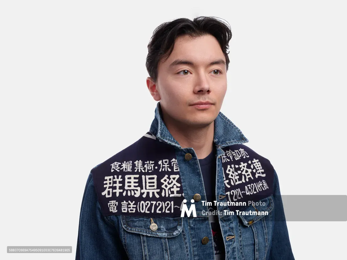 A young Asian man gazes directly at the camera with a composed, contemplative expression against a pristine white backdrop. He wears a distinctive denim jacket adorned with white Japanese characters and telephone numbers embroidered across the chest and sleeve, suggesting vintage workwear or business advertising. The even studio lighting creates soft shadows that define his features while maintaining the jacket's rich blue texture and intricate stitching details.