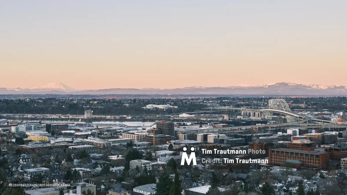 The sprawling neighborhoods of Northwest Portland unfold beneath a pastel dusk sky, where residential homes nestle between modern apartment complexes and commercial buildings. The iconic white arches of the Fremont Bridge span the distant Willamette River, while the snow-capped silhouette of Mount Saint Helens anchors the horizon in Washington State. Golden hour light bathes the urban landscape in warm tones, creating a serene contrast between the intimate scale of residential streets and the grandeur of the Cascade Range beyond.