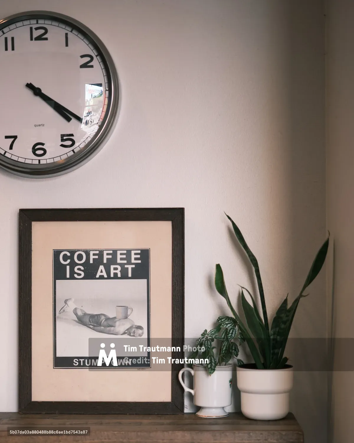 A wall clock, framed 'Coffee is Art' poster from Stumptown coffee shop, and potted plants arranged on a wooden shelf against a beige wall. The scene captures the cozy, artistic atmosphere of the Portland coffee shop interior.