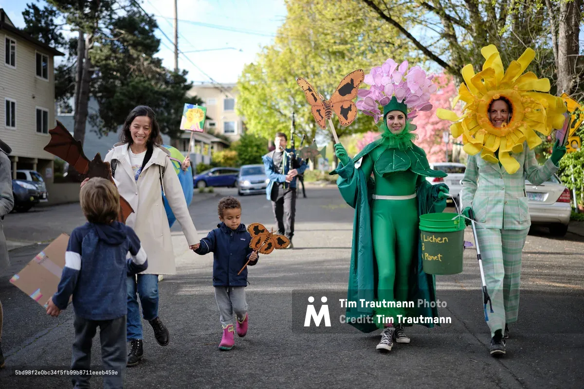A group of people in colorful environmental-themed costumes including a large green leaf character and yellow sunflower walk down a residential street in Portland's Central Eastside district. Children and adults participate in what appears to be a community parade or environmental awareness event, with some carrying butterfly decorations and eco-themed props.