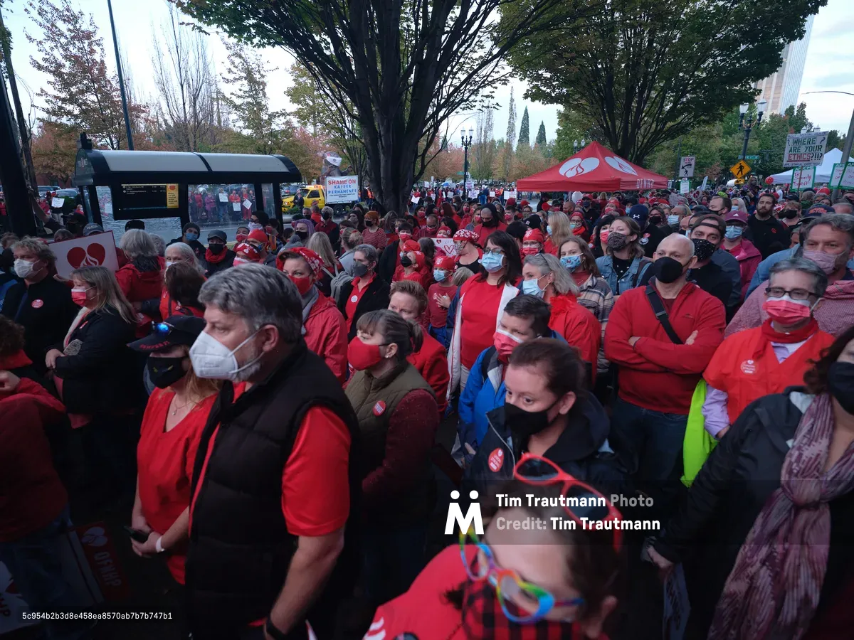 Hundreds of Kaiser Permanente nurses flood the plaza near the healthcare giant's Portland tower, a crimson tide of solidarity beneath autumn's canopy. Face masks create a mosaic of protection and protest as healthcare workers stand shoulder-to-shoulder in coordinated red attire, their unity palpable in the crisp Oregon air. The urban forest of downtown Portland frames this moment of labor action, where professional dedication meets collective resolve. Red popup tents and protest signs punctuate the crowd, while the city's skyline bears witness to this powerful display of healthcare worker solidarity.
