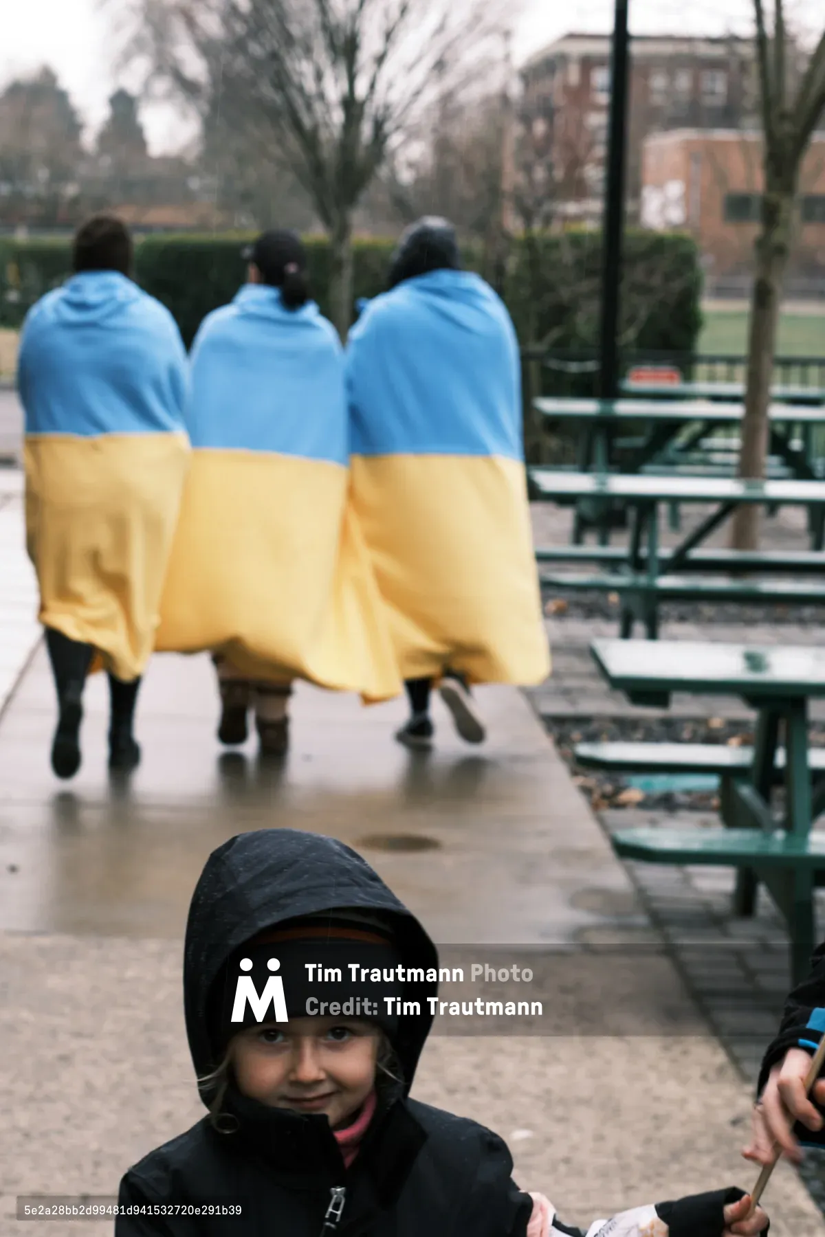 Three figures draped in Ukrainian flags stand with their backs turned on the rain-slicked plaza outside Portland's Revolution Hall, their blue and yellow shrouds creating a haunting tableau of solidarity. A young child in a dark hooded jacket sits in the foreground, gazing directly at the camera with knowing eyes that belie their years. The overcast February sky and bare winter trees frame this prescient moment of protest, captured in the days before Russian forces would cross into Ukraine.