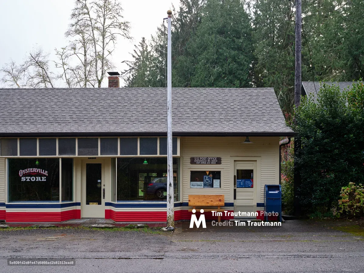 A weathered cream-colored building houses both the Oysterville Store and United States Post Office along a quiet stretch of Oysterville Road in Pacific County, Washington. The modest structure features patriotic red, white, and blue trim beneath gray shingles, with large storefront windows reflecting the overcast Pacific Northwest sky. A solitary wooden bench sits beneath the post office window, while a blue mailbox stands sentinel beside the entrance, embodying the unhurried pace of this historic oyster farming community on the Long Beach Peninsula.