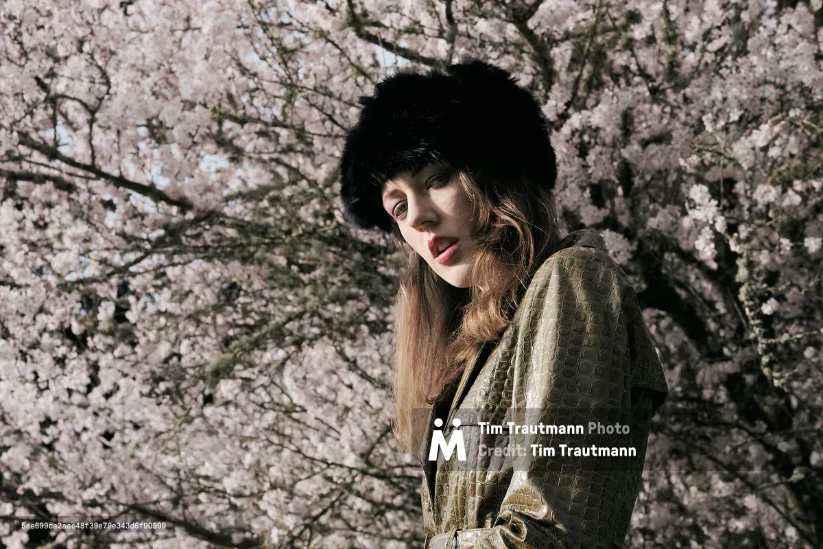 A portrait of Rhianna Atwood standing in front of a fully bloomed cherry blossom tree, its soft pink flowers filling the background. She wears an olive green crocodile-textured leather coat and a black fluffy fur hat, with long brown hair resting over her shoulders. She gazes downward with a serene expression in warm natural sunlight.