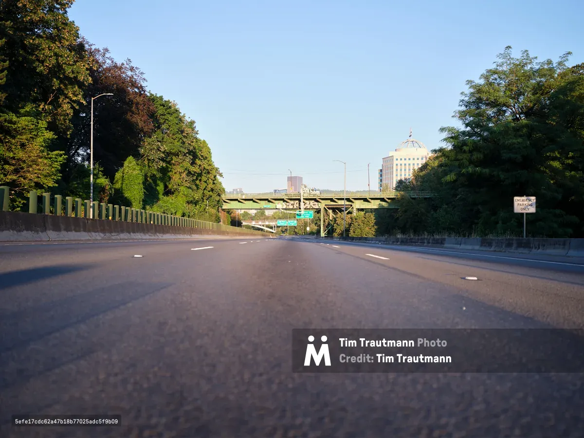 A low-angle view of the empty Banfield Freeway (I-84) in Portland's Lloyd District, showing the asphalt road surface with lane markings leading toward downtown buildings visible in the distance. Lush green trees line both sides of the highway, with a concrete overpass bridge crossing above and clear blue skies overhead.