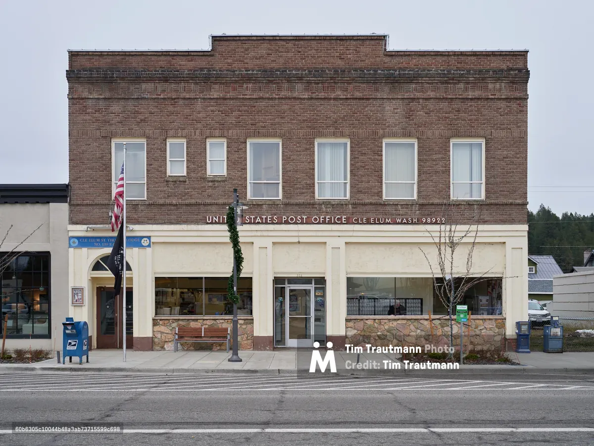 A weathered brick United States Post Office stands resolute along East 1st Street in Cle Elum, Washington, its classic early 20th-century facade crowned with traditional red brick and cream-colored stone trim. The overcast Pacific Northwest sky casts even light across the building's seven tall windows, while the American flag hangs quietly beside the entrance. Stone wainscoting and large storefront windows reveal the building's dual role as both federal facility and community anchor, set against the backdrop of the Cascade Mountains' forested hills.