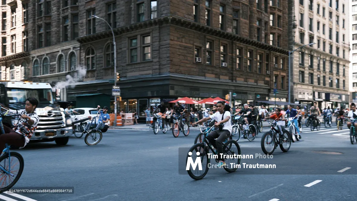 A dynamic street scene captures dozens of cyclists flowing through the intersection at 7th Avenue in Manhattan's Garment District. The afternoon light casts long shadows across the asphalt as riders on various bicycles navigate between yellow cabs and commercial vehicles. Historic stone facades with ornate cornices frame the urban tableau, while red umbrellas from a sidewalk vendor add splashes of color to the predominantly neutral palette of weathered limestone and concrete.