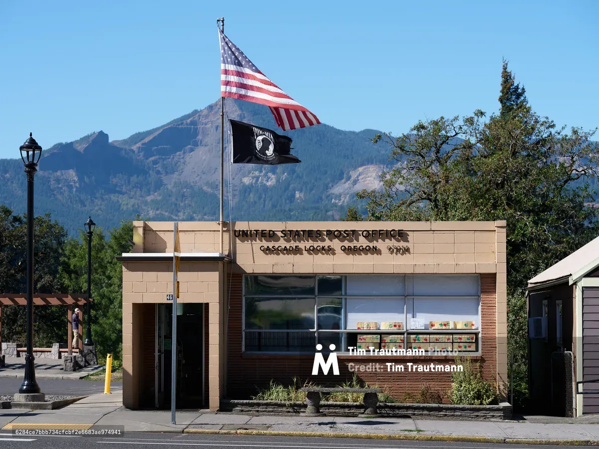 The mid-century brick and concrete United States Post Office stands sentinel in Cascade Locks, Oregon, its modernist lines softened by the dramatic backdrop of forested mountains rising into crystal blue sky. Two flags—the Stars and Stripes and a black POW/MIA banner—flutter from a single pole, while vintage street lamps frame the humble federal building. The Columbia River Gorge's towering peaks create a theatrical backdrop for this essential piece of small-town American infrastructure, where community connection meets governmental service.