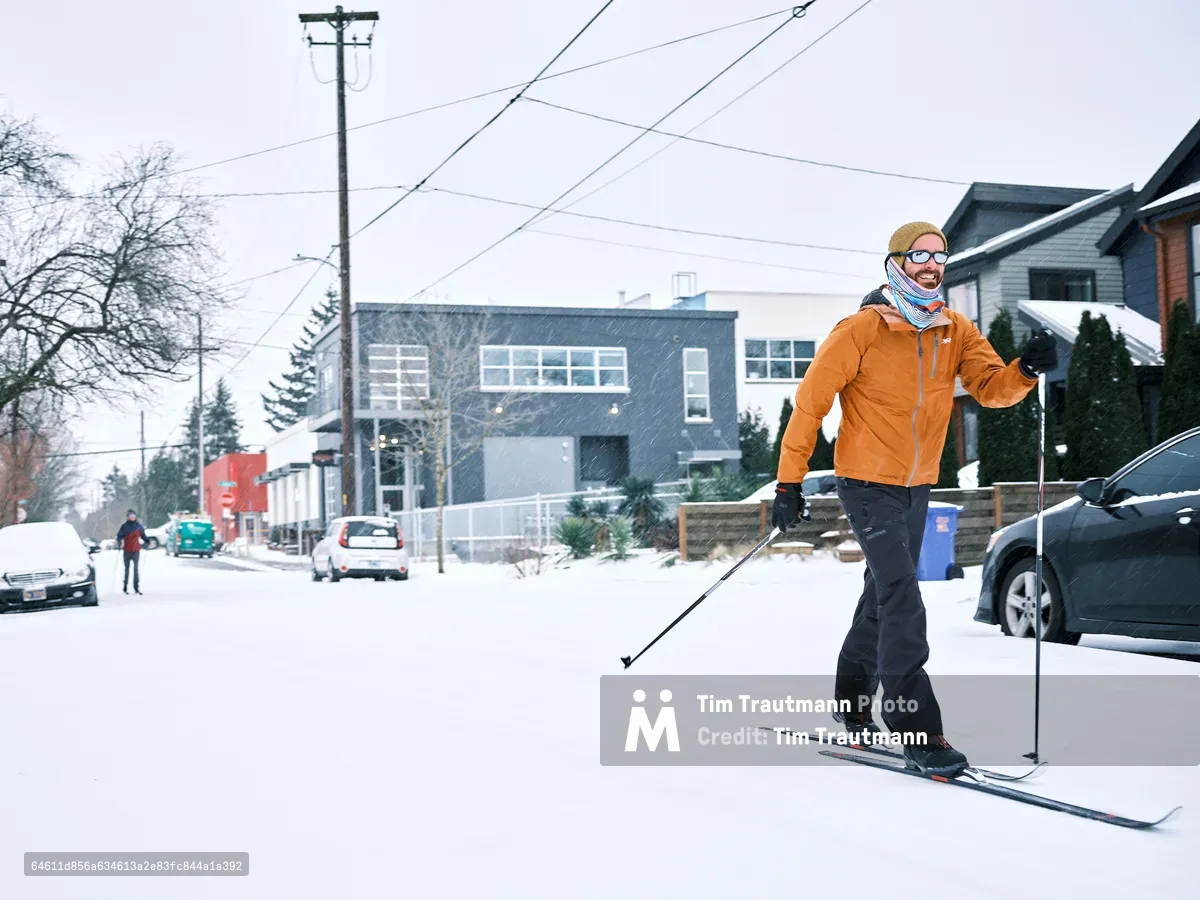 A man cross-country skis down a snow-covered residential street in Portland's Alberta Arts District during a winter snowstorm. He wears an orange jacket, black ski pants, a mustard yellow beanie, and mirrored sunglasses, smiling as he glides past parked cars. A second skier in a red jacket is visible further down the street in the background, along with modern homes, utility poles, and lightly falling snow.