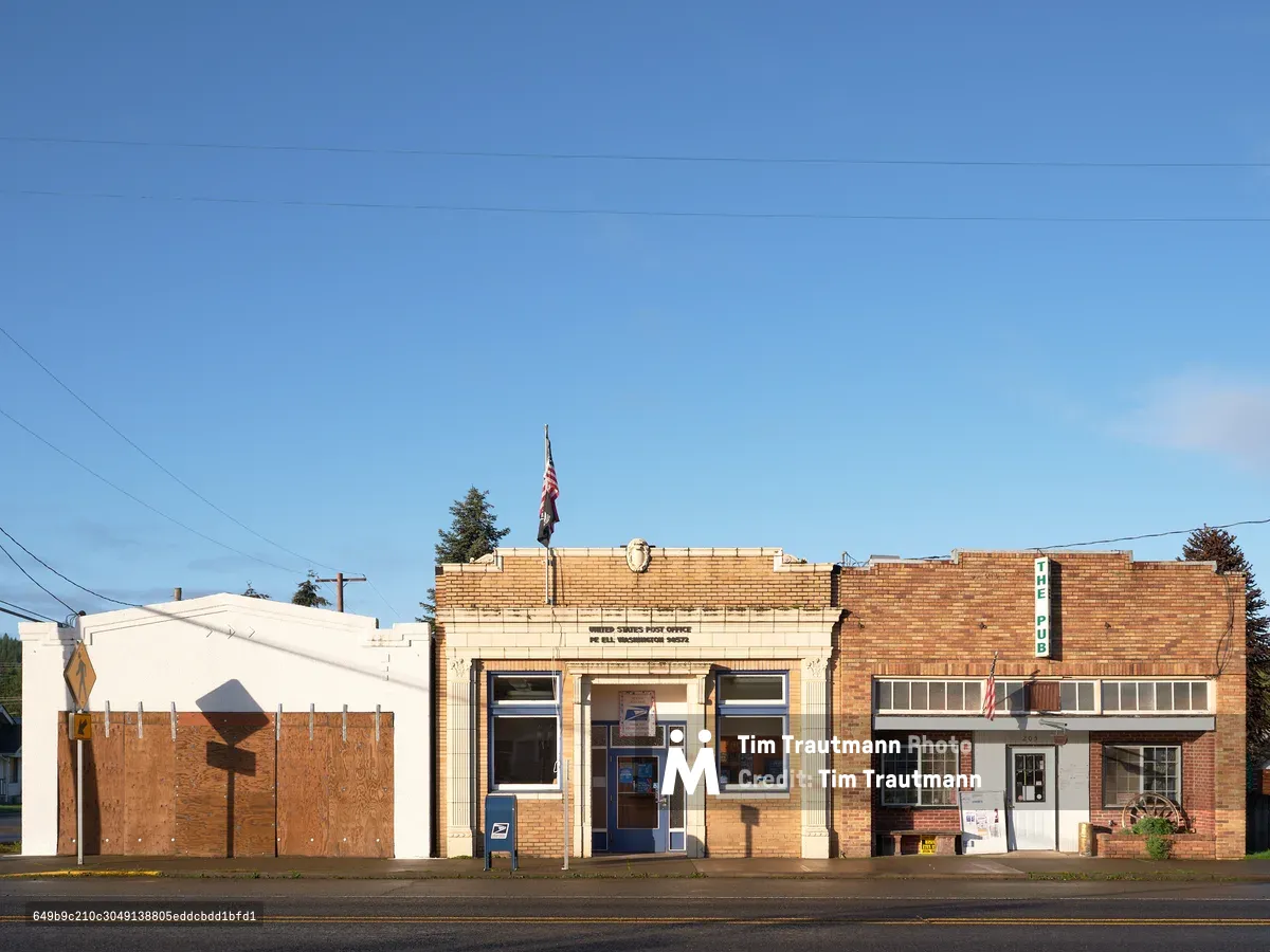 The modest brick United States Post Office stands sentinel along Main Street in Pe Ell, Washington, its cream-colored facade crowned by an American flag against an expansive cerulean sky. The small-town postal building anchors a row of weathered brick storefronts, their varied textures telling stories of rural resilience under the clear Pacific Northwest light. Power lines sketch geometric patterns overhead while shadows stretch across the quiet street, capturing the unhurried rhythm of this Lewis County community.
