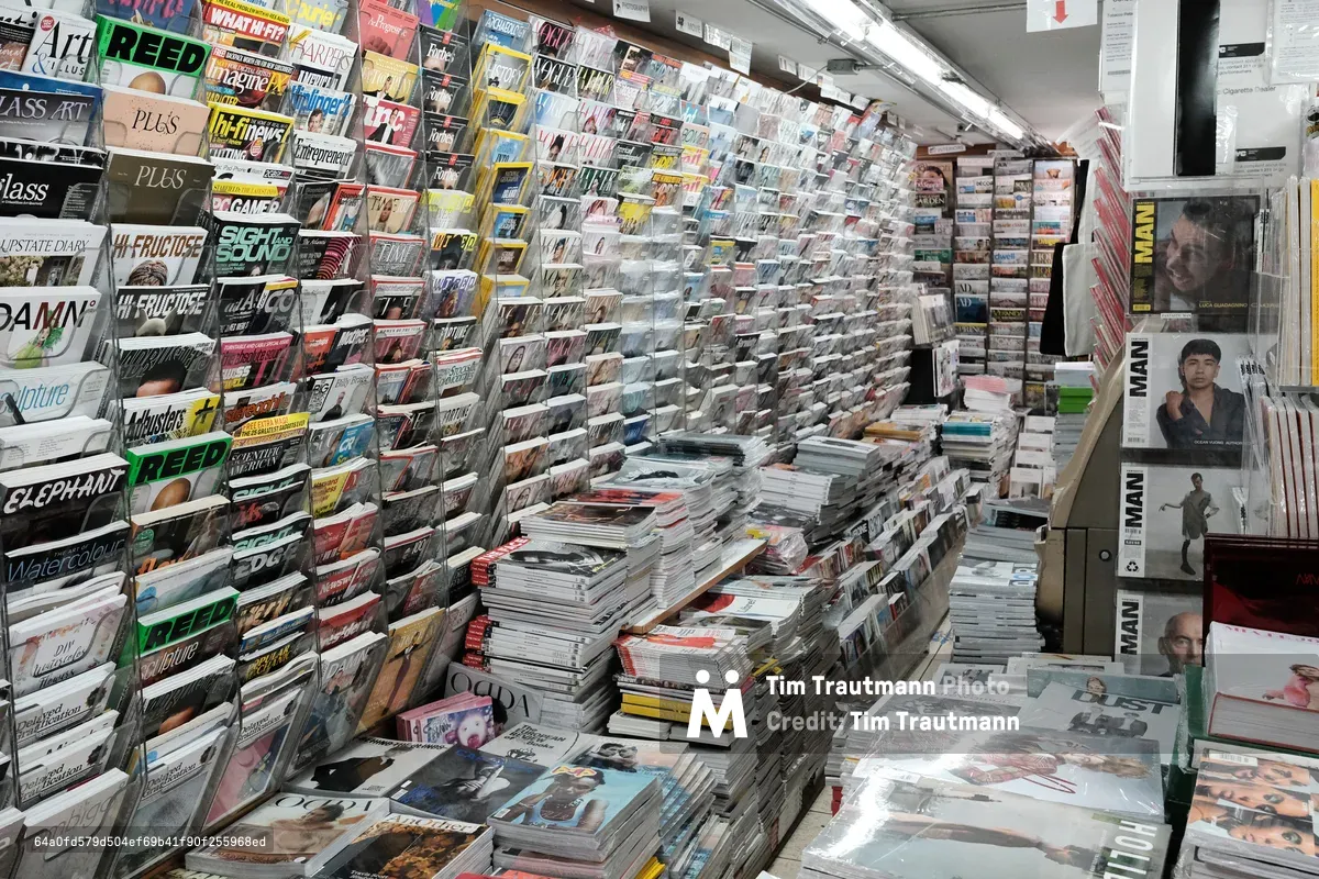 The cramped interior of a traditional newsstand on Prince Street in SoHo reveals an overwhelming kaleidoscope of publications. Floor-to-ceiling magazine racks create narrow corridors lined with hundreds of titles, from fashion glossies to niche periodicals, while harsh fluorescent lighting illuminates the densely packed space. Stacks of fresh magazines and newspapers sprawl across every available surface, creating a labyrinthine environment that speaks to the enduring appetite for print media in Manhattan's cultural heart.