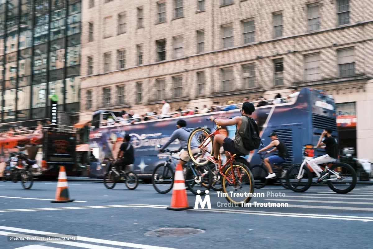 Motion blur captures the kinetic energy of cyclists racing down Manhattan's Seventh Avenue, their figures streaking past in a symphony of movement against the stoic backdrop of brick tenements and modern glass facades. The photographer's intentional camera movement transforms the urban thoroughfare into a river of color and speed, where orange traffic cones punctuate the asphalt like sentinels guiding the flow of human-powered transit. The contrast between the sharp architectural details and the fluid cyclists creates a visceral sense of metropolitan rhythm and the relentless pulse of New York City street life.