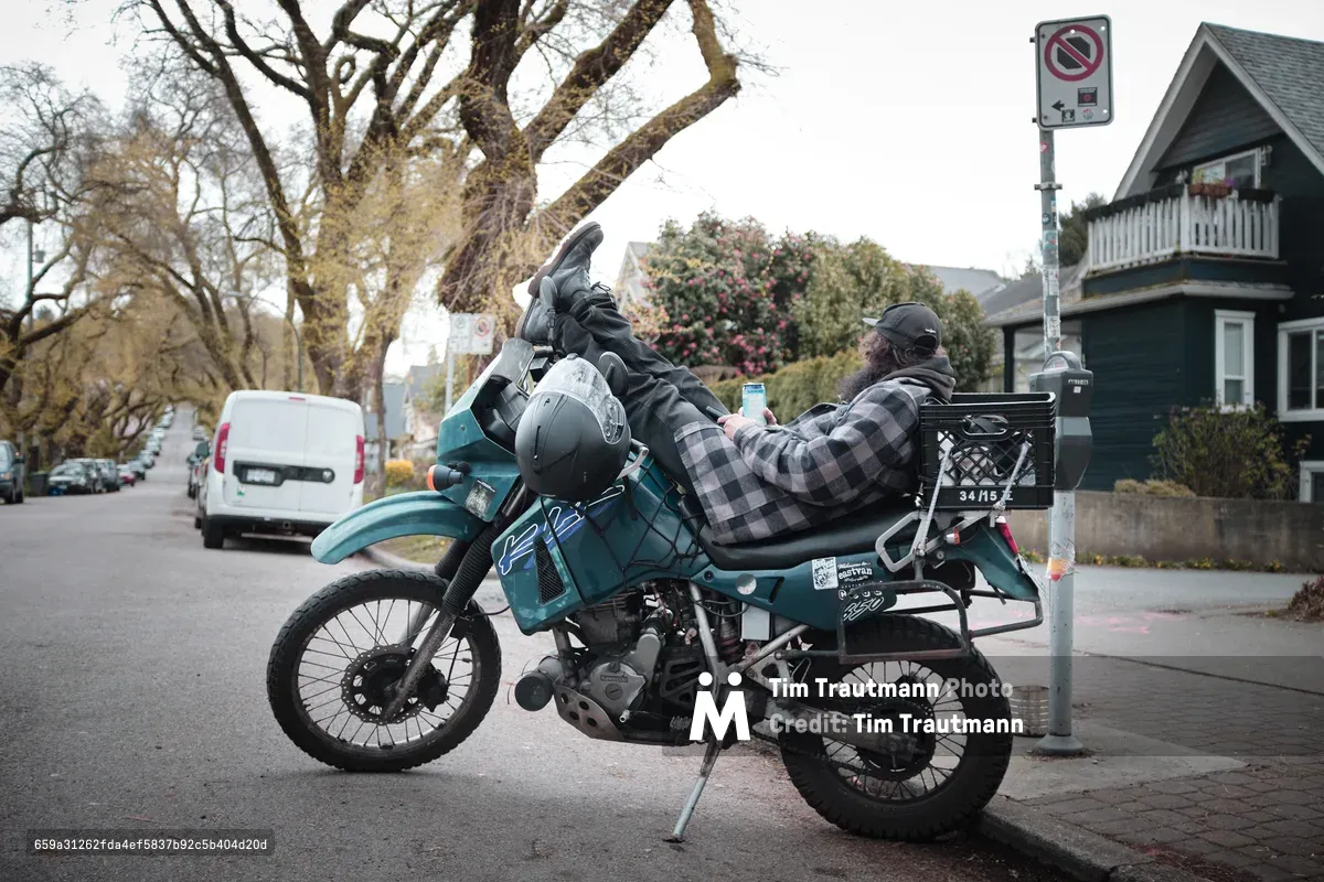A weathered adventure motorcycle loaded with travel gear sits parked on East 6th Avenue in Vancouver's Grandview-Woodland neighborhood, its rider taking a contemplative break. The teal dual-sport bike, adorned with scratches and travel stickers, bears witness to countless miles beneath the bare branches of winter trees. Residential charm surrounds the scene with heritage homes and urban signage creating an atmospheric backdrop under overcast skies.