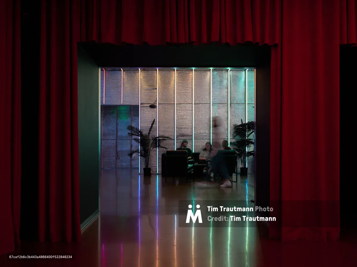 View through red curtains into a contemporary restaurant dining room featuring vertical neon light strips in rainbow colors along wood-paneled walls, with diners seated at tables and palm plants as decor. The polished floor reflects the vibrant lighting creating a modern, upscale atmosphere.