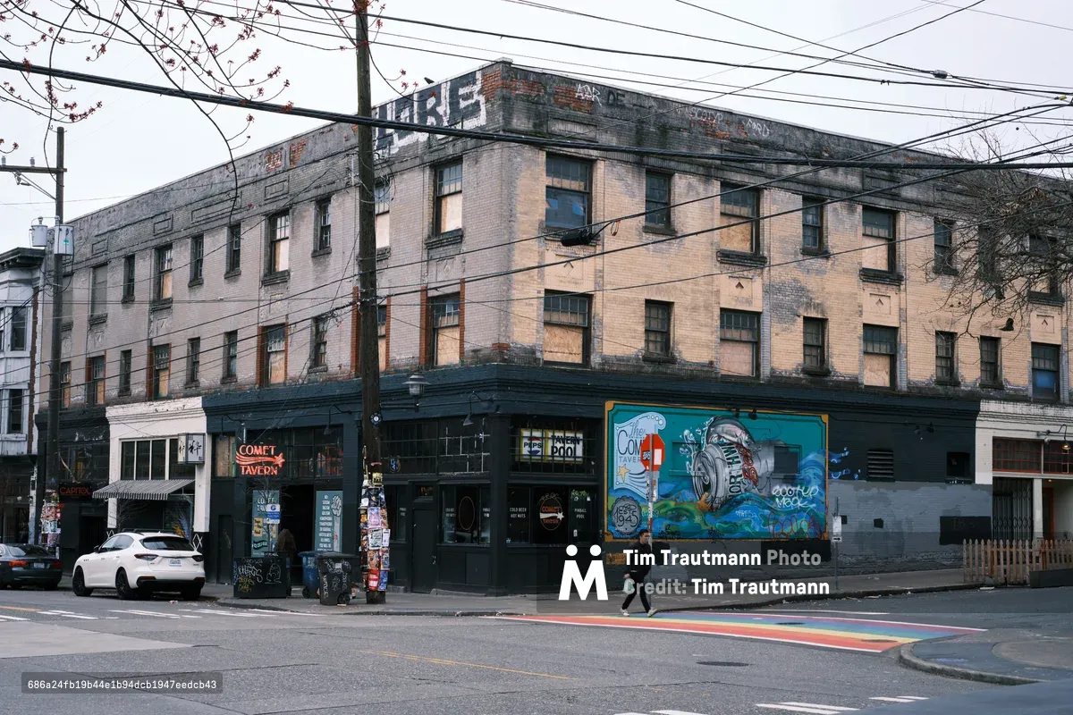 The Comet Tavern anchors a weathered brick corner building in Seattle's Capitol Hill neighborhood, where power lines slice through an overcast sky above rain-slicked streets. The building's faded industrial facade tells stories of urban evolution, with peeling paint and graffiti creating a textural palimpsest against warm brick tones. A solitary figure crosses the intersection marked by rainbow crosswalk stripes, while vintage neon signs and vibrant street murals inject color into the moody Pacific Northwest atmosphere.