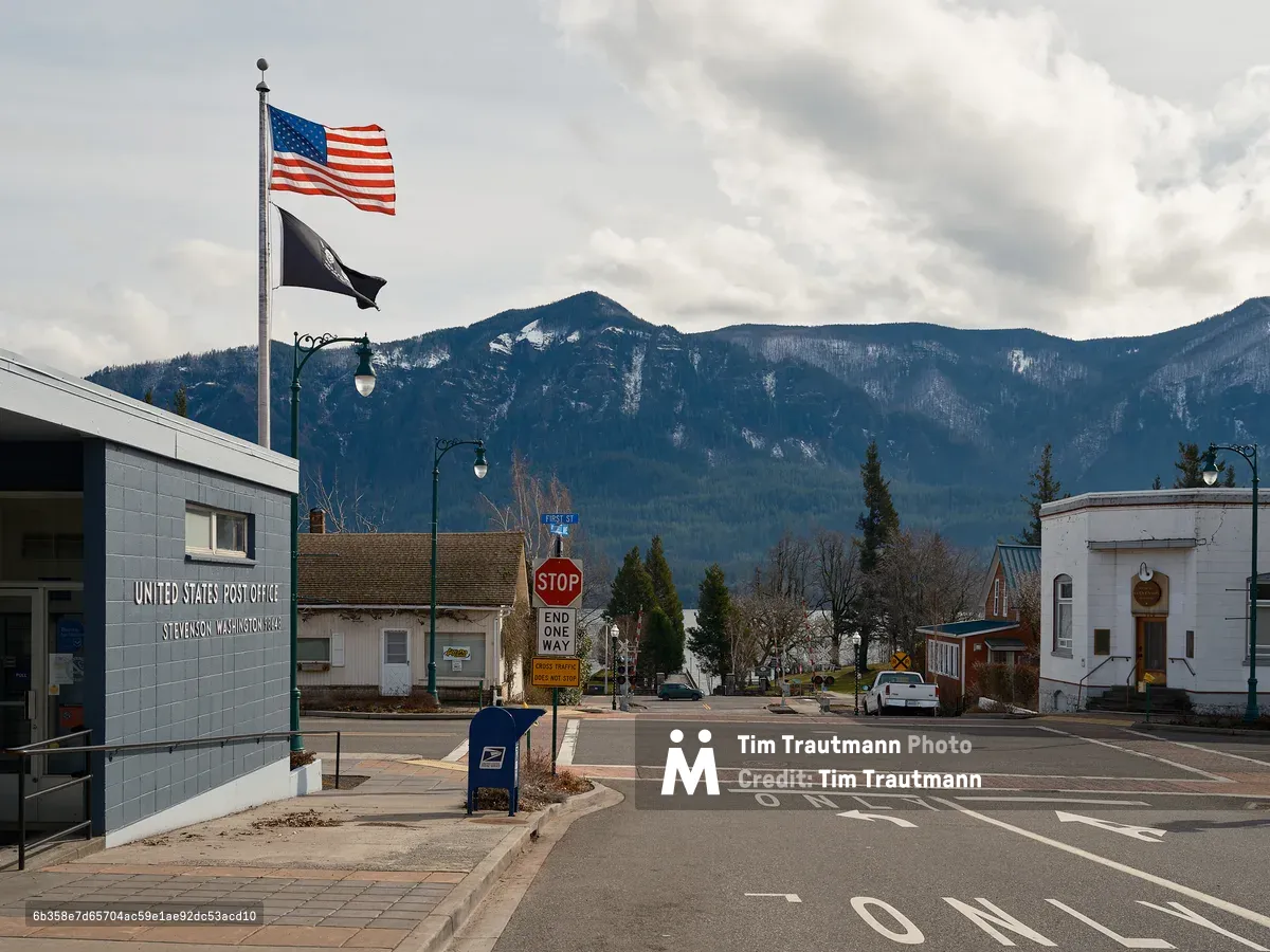 The United States Post Office in Stevenson, Washington, a single-story blue brick building with signage reading "Stevenson Washington 98648." An American flag and a black POW/MIA flag fly from a tall flagpole. A blue USPS mailbox sits on the sidewalk near a stop sign and "End One Way" sign at an intersection. In the background, a quiet small-town main street leads toward the Columbia River Gorge, with densely forested mountains dusted with snow rising dramatically under a partly cloudy sky.