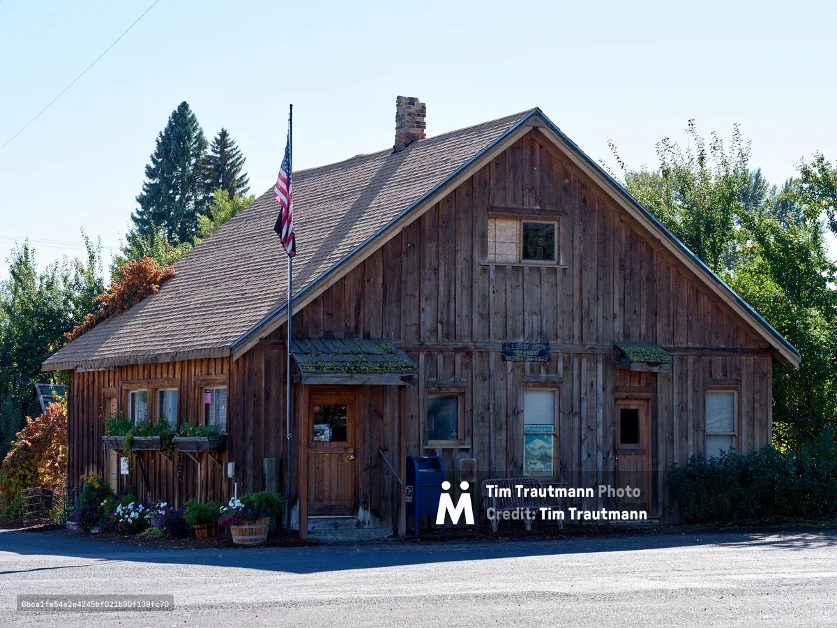 A weathered wooden post office building stands as a testament to small-town America in Glenwood, Washington. The rustic cedar-shingled structure features moss-covered eaves and a prominent American flag, creating an atmospheric portrait of rural postal service. Soft evening light bathes the scene, highlighting the building's authentic patina and the carefully tended flower boxes that add touches of color to the weathered facade. The composition captures both the utilitarian purpose and enduring charm of this community cornerstone nestled among towering evergreens.