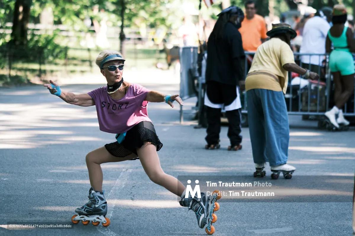 A joyful woman in purple athletic wear and sunglasses glides through Central Park on inline skates, her arms spread wide in triumphant celebration. Dappled sunlight filters through the leafy canopy above, creating a warm, golden atmosphere on the park's paved pathways. Behind her, other park visitors stroll leisurely, creating a perfect contrast between her dynamic energy and the peaceful weekend ambiance. The composition captures the pure freedom and exhilaration of urban recreation in Manhattan's beloved green oasis.
