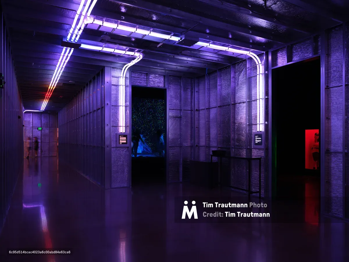 A dramatic interior space at Hopscotch in Portland, Oregon featuring purple and blue neon lighting, concrete walls, and reflective floors creating a cyberpunk-inspired atmosphere. The space includes curved neon tubes on the ceiling, display screens, and red accent lighting along dark corridors.