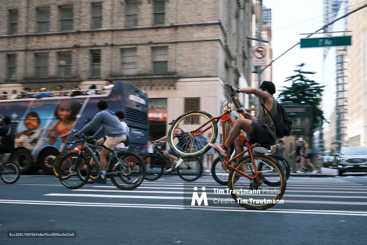 A young cyclist defies gravity, performing a dramatic wheelie on his orange BMX bike as he crosses a busy Manhattan intersection. The motion-blurred scene captures the raw energy of urban street culture, with a city bus and fellow riders creating a dynamic backdrop against the towering brick facades of Midtown. Golden hour light bathes the asphalt, transforming an ordinary crosswalk into a stage for spontaneous street performance.