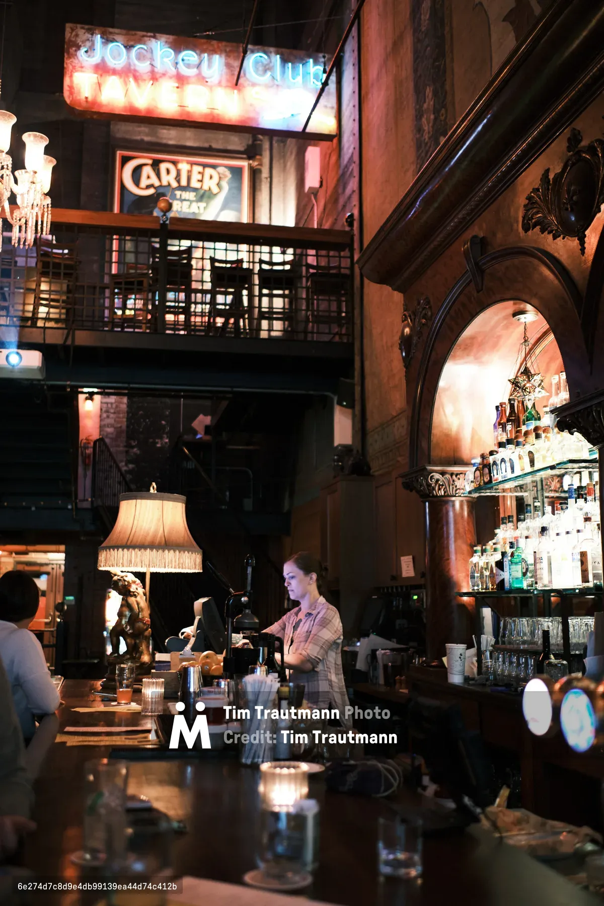A bartender works behind the mahogany bar at Portland's historic Backstage Bar, illuminated by the warm glow of vintage neon signs reading 'Jockey Club' and 'Carter's The Great.' The atmospheric interior features soaring Gothic arches, ornate moldings, and a mezzanine level with iron railings, while fringed table lamps and carefully arranged bottles create theatrical shadows. The composition captures the establishment's vaudeville heritage through its dramatic lighting and layered architectural details.