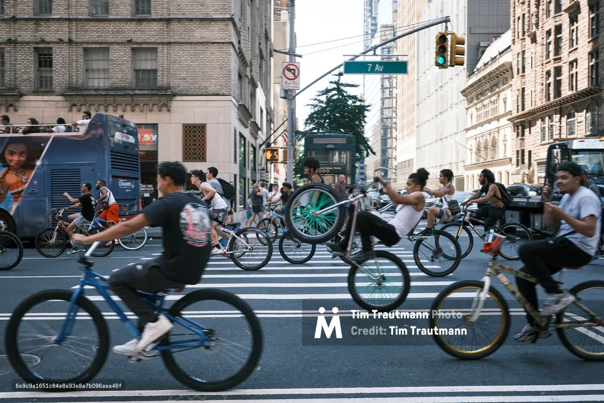 A dynamic swarm of cyclists floods 7th Avenue in Manhattan during what appears to be a Critical Mass ride, their bicycles creating a kinetic blur of motion against the towering urban canyon. The late afternoon light catches the handlebars and spokes as dozens of riders pedal through the intersection, transforming the typically car-dominated thoroughfare into a river of human-powered transportation. Classical pre-war architecture rises on both sides, with ornate facades and fire escapes framing this moment of collective urban rebellion.