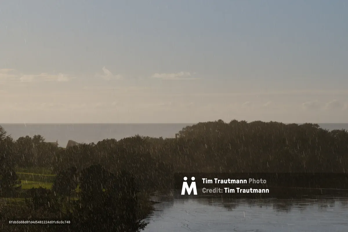 A dramatic scene of intense rainfall falling over a forested coastal landscape in Gleneden Beach, Oregon, with visible rain streaks across the frame, dark storm clouds overhead, and a body of water in the foreground reflecting the moody sky.