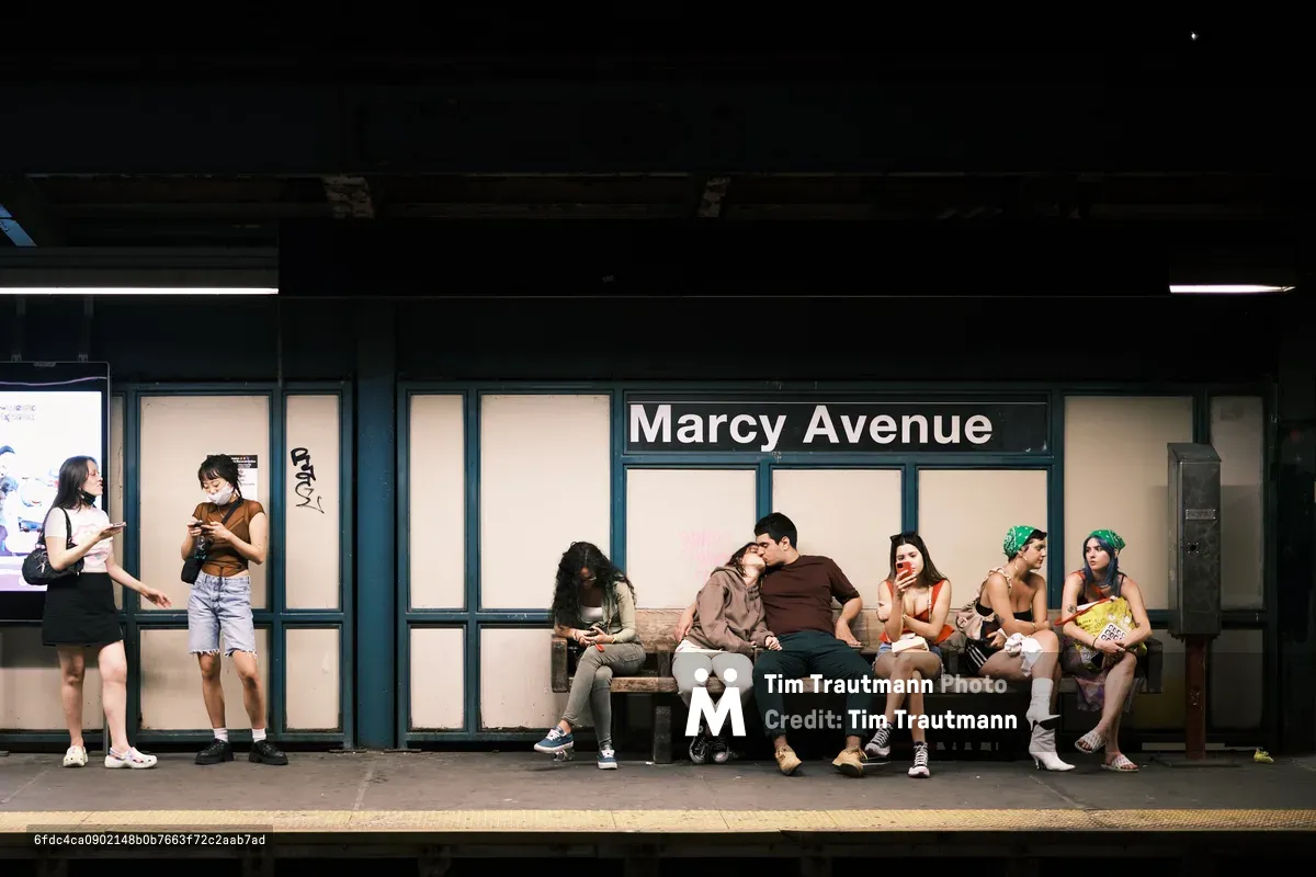 Passengers wait beneath the stark fluorescent lighting of Brooklyn's Marcy Avenue subway platform, their bodies creating a intimate tableau against the institutional green tiling. A couple shares a tender moment in the center of the bench while fellow commuters engage with their phones and conversations, each absorbed in their own urban rituals. The warm summer evening light filters through the station's windows, softening the harsh underground environment and illuminating this slice of contemporary New York transit life.