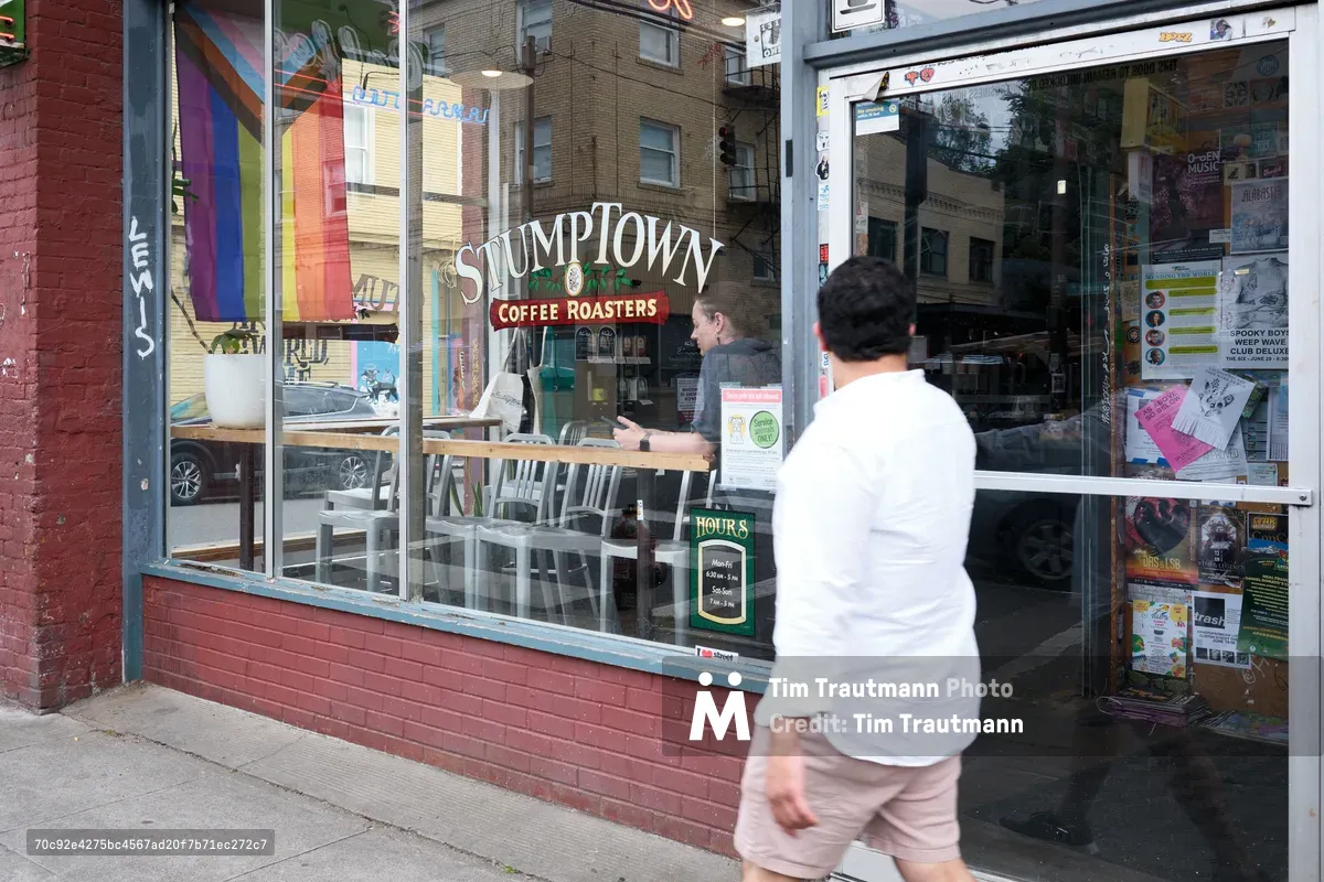 A person in a white shirt walks past the storefront window of Stumptown Coffee Roasters on Southeast Belmont Street in Portland's Sunnyside neighborhood. The large windows display the coffee shop's interior with customers seated at tables, while colorful flyers and community notices are posted on the glass.