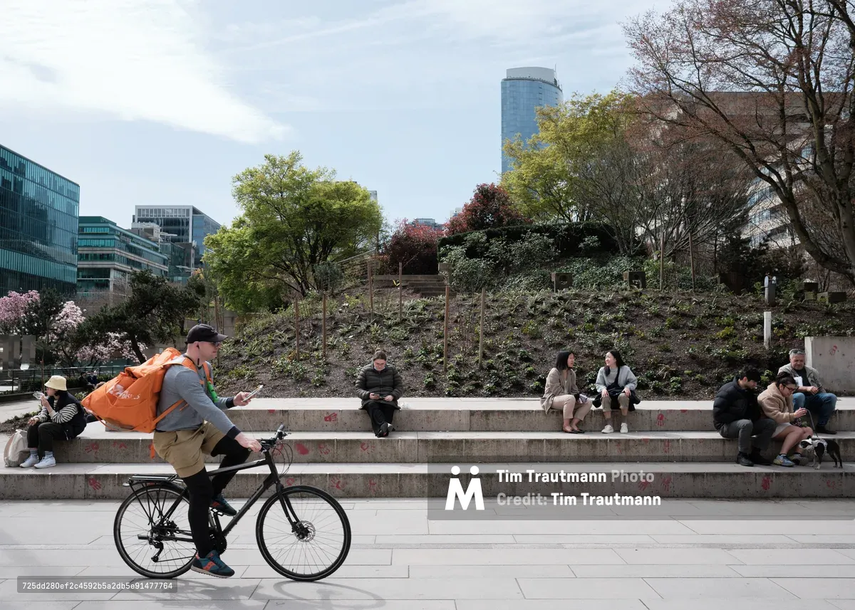 A delivery cyclist in a vibrant orange jacket glides past tiered concrete steps where young people gather in casual clusters beneath spring's awakening foliage. The scene unfolds against Vancouver's distinctive downtown skyline, with glass towers rising behind a hillside garden showing the tender greens and burgundy blossoms of the Pacific Northwest's seasonal transition. Soft overcast light bathes the urban plaza, creating an intimate tableau of city life in motion.