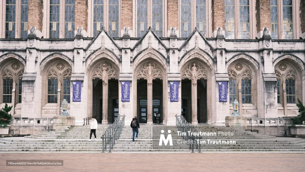The imposing Gothic Revival facade of Suzzallo Library dominates the frame with its soaring pointed arches and ornate stonework, crowned by rows of tall mullioned windows that catch the soft daylight. Purple welcome banners flutter between the carved columns, while two solitary figures navigate the broad stone steps, dwarfed by the architectural monumentality. The warm-toned limestone contrasts beautifully with the deep shadows cast by the elaborate tracery, creating a scene that balances academic solemnity with accessible human scale.