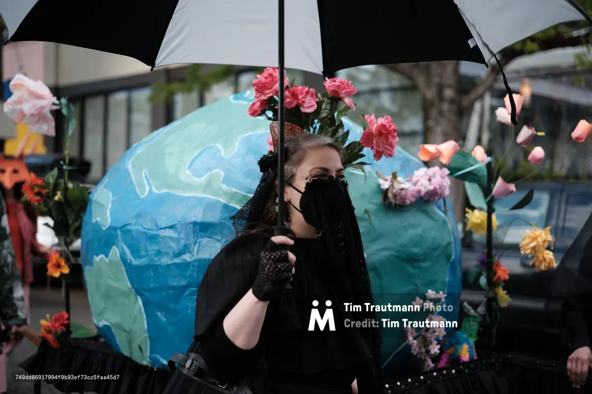 A person wearing all black with a face mask stands under a black and white umbrella, adorned with a large blue and green Earth costume decorated with colorful flowers. The scene appears to be at an outdoor environmental protest or demonstration in Portland, Oregon.