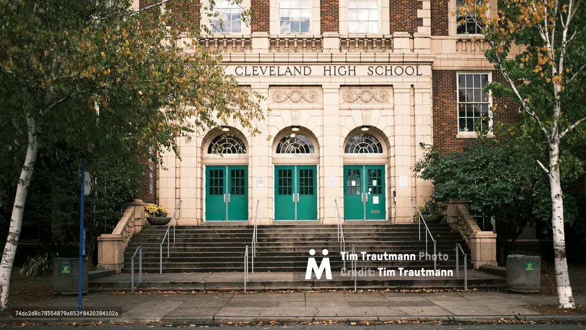 The stately historic facade of Grover Cleveland High School in Portland, Oregon, photographed on an autumn day. The building features a classical red brick and cream stone exterior with three arched entryways fitted with teal green double doors and fanlight windows above. "Cleveland High School" is carved in stone above the central entrance. Wide stone steps with metal handrails lead up to the three entrances. White-barked birch trees with yellowing autumn leaves frame the building on both sides, and fallen leaves scatter the sidewalk in the foreground.