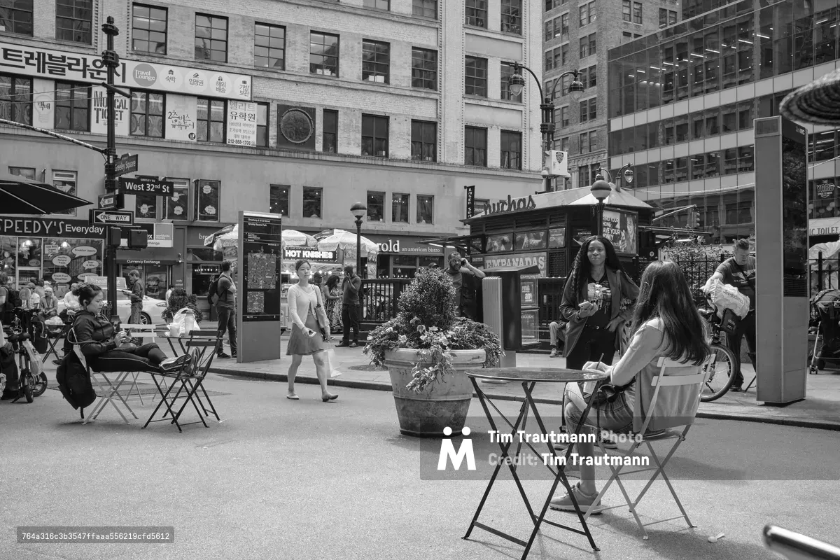 In the bustling heart of Herald Square, Manhattan, pedestrians claim moments of urban tranquility at scattered café tables amid the commercial chaos. The monochromatic composition captures the rhythmic dance of city life, where solitary figures pause with coffee and conversation against a backdrop of towering brick facades and Korean signage. Geometric folding chairs and planters create intimate pockets of respite, while streams of shoppers navigate between the outdoor seating and storefront displays.
