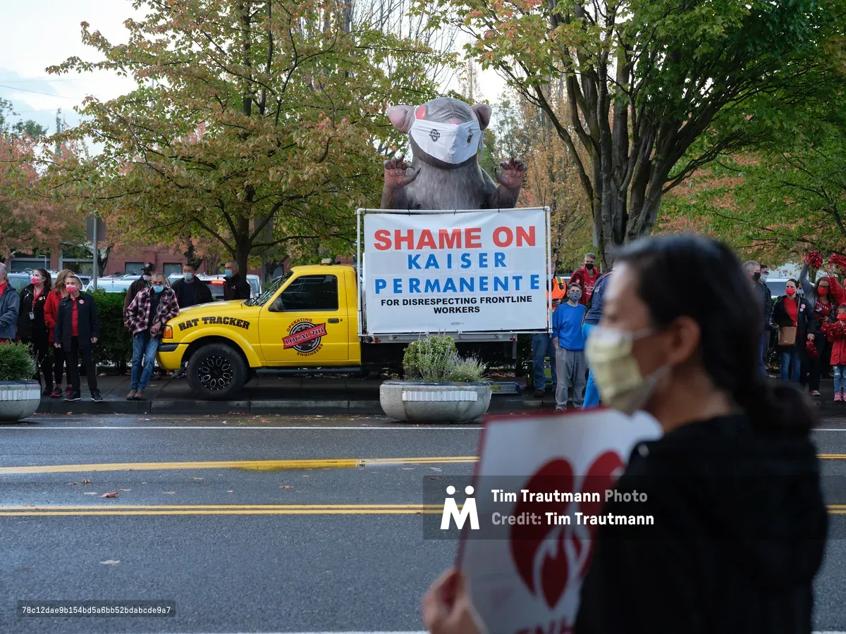 A towering inflatable rat protest figure dominates the scene outside Kaiser Permanente Tower in Portland's Lloyd District, its menacing presence amplified by autumn's golden canopy. Red-clad nurses and supporters gather in solidarity along Northeast Multnomah Street, their masked faces reflecting both pandemic precautions and labor tensions. The bold white protest sign declaring 'SHAME ON KAISER PERMANENTE' cuts through the overcast afternoon light, while a yellow Rat Tracker truck anchors the demonstration with industrial determination.