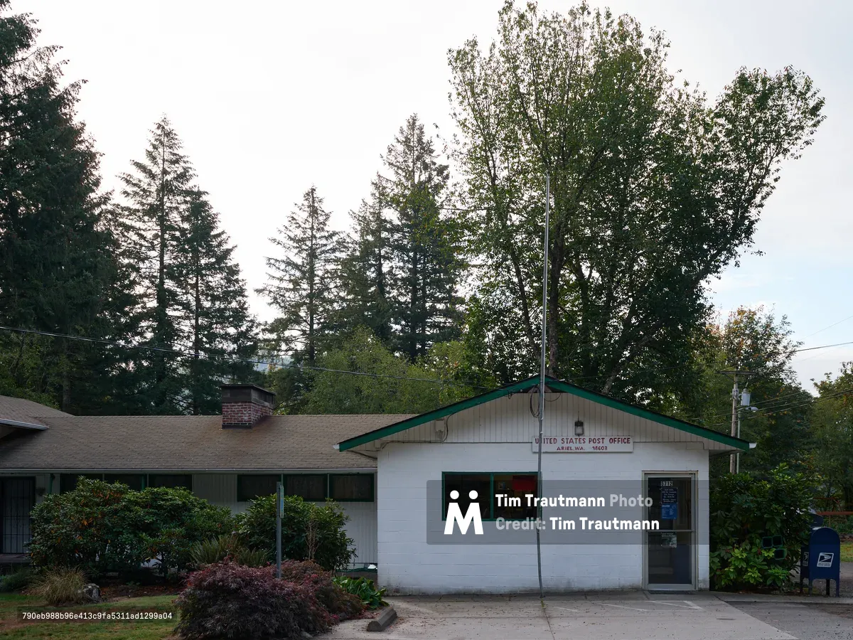 A modest white clapboard post office building sits beneath towering evergreens in the rural community of Ariel, Washington. The structure features a distinctive green metal roof and trim, with the official 'United States Post Office' signage proudly displayed above the entrance. Mature Pacific Northwest conifers create a natural canopy overhead, while a blue USPS collection box stands sentinel beside the front door, embodying the enduring connection between federal services and small American towns.